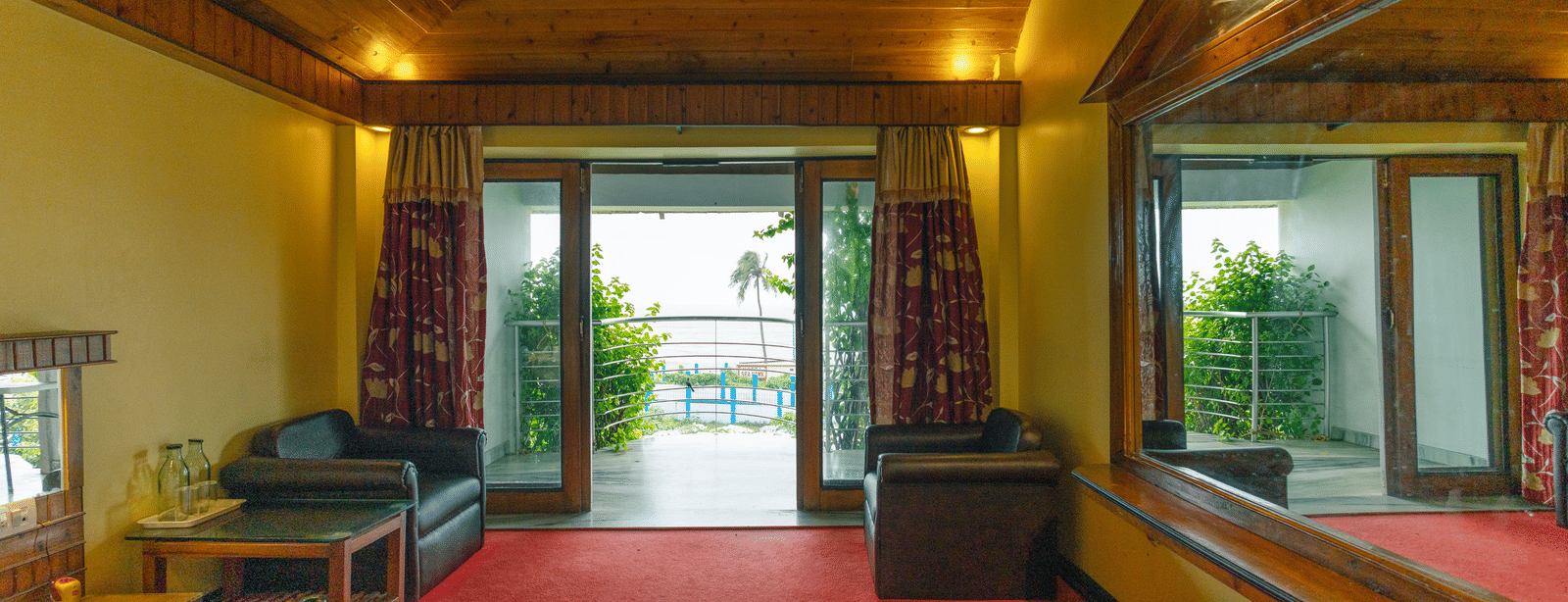 A bedroom with large windows leading to a balcony, 2 armchairs, a table, a mirror, wooden ceiling, and a king size bed at Hotel Seagull Digha.